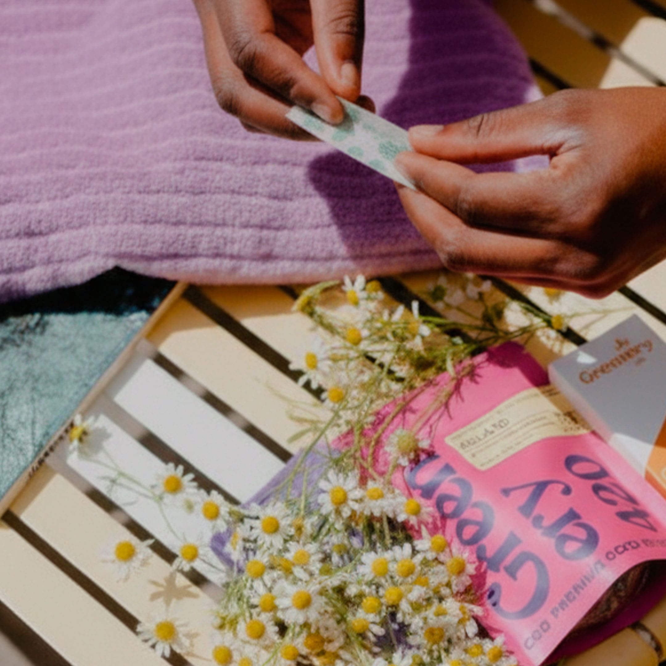La Manzanilla cómo alternativa al tabaco Manos enrollando papel de fumar junto a flores de manzanilla y un paquete de Greenery CBD sobre una superficie iluminada por el sol.